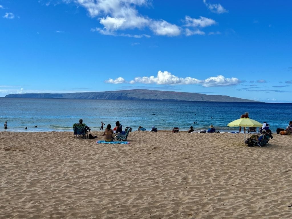 People relaxing on a wide sandy beach with clear skies and an island seen in the distance - beaches and water activities are some of the best things to do in Kihei