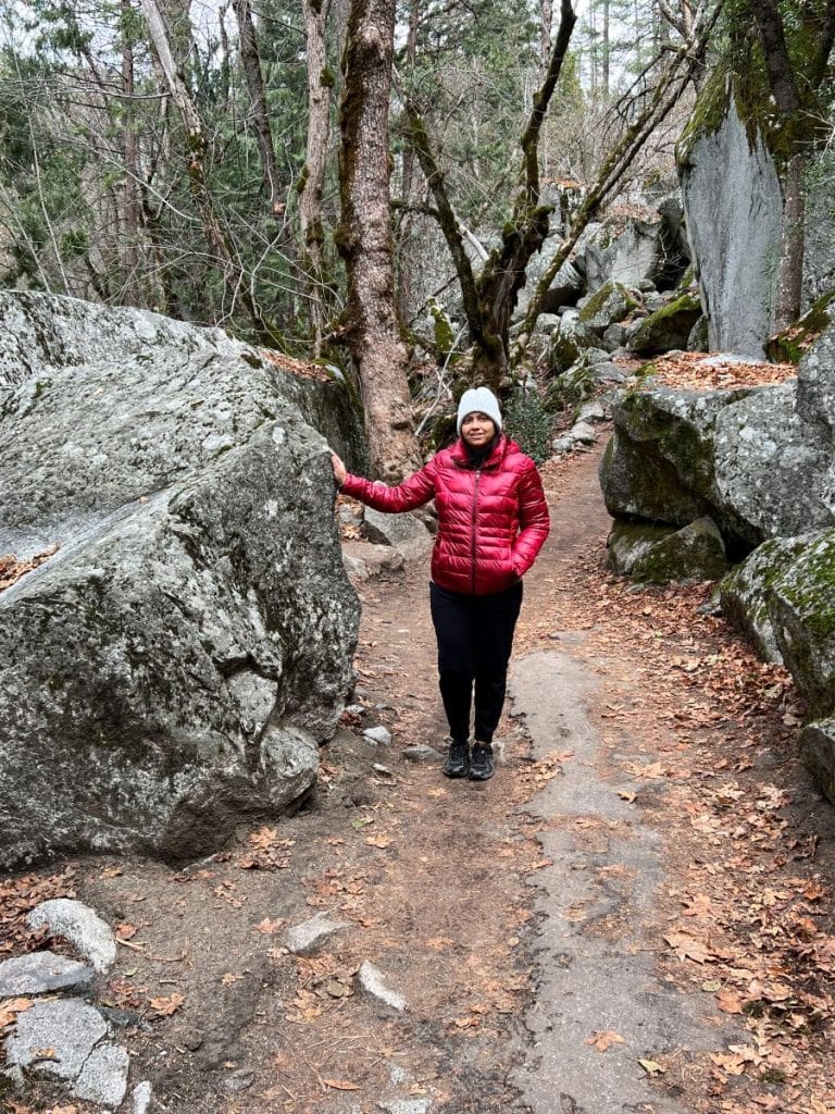 Girl in a red jacket posing on a hiking trail by standing next to a big boulder.