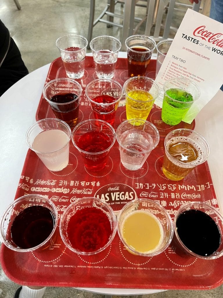 A red try with 16 clear glasses with colorful cola-cola flavors