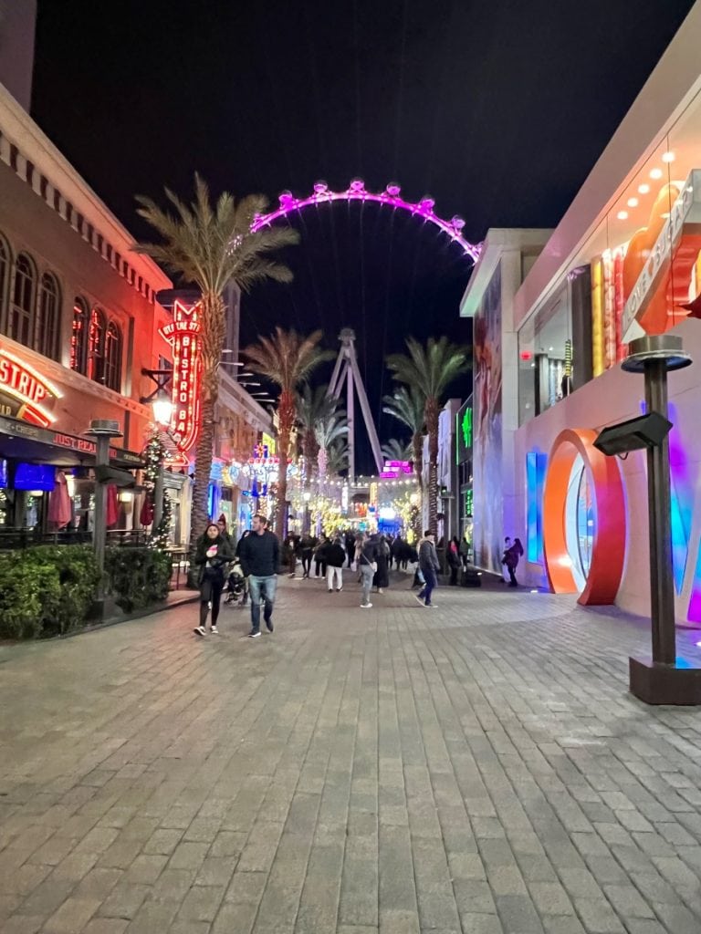 A street at night lined with colorful shops on both sides and the High Roller observatory wheel in the background