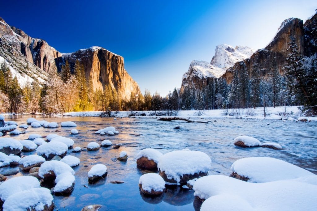 Merced River in Yosemite during winter with snow on boulders and rocks. Granite mountains in the distance have snow on them
